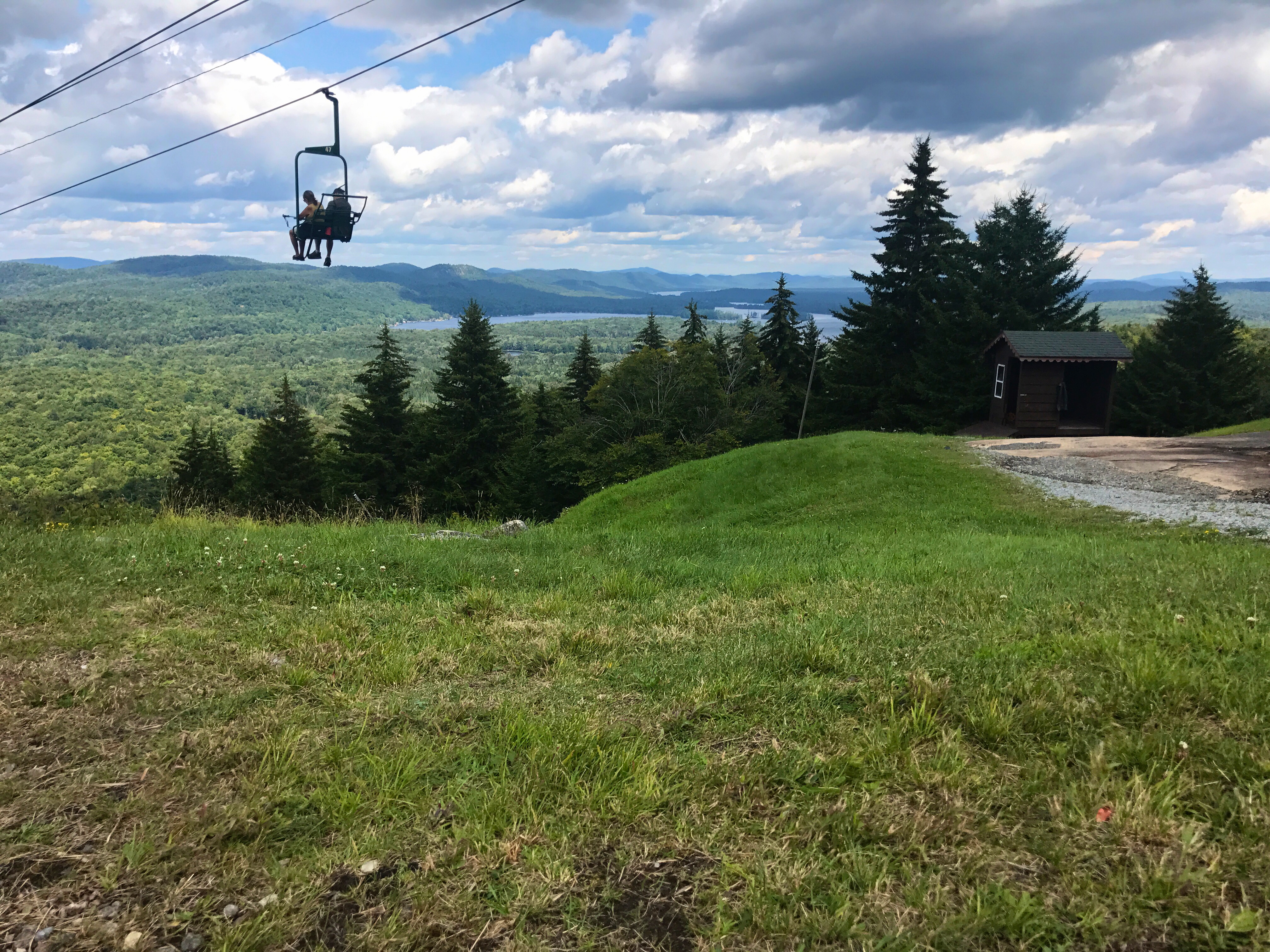 View from McCauley Mountain. The picture portrays two people enjoying a ride on the chair lift located at McCauley Mountain, and a view of the mountains, trees, and lakes.